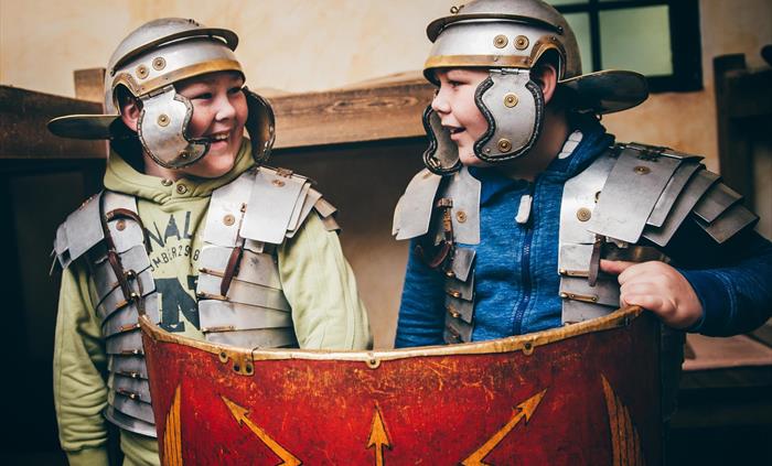 Two young boys wearing Roman helmets and armour. They are holding up a red Roman shield and smiling.
